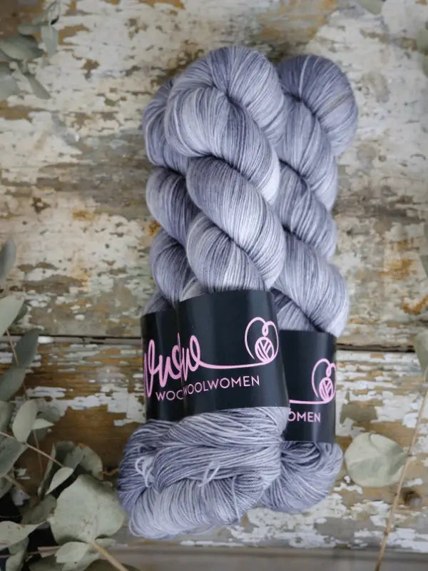 Two skeins of WoolWomen Merino Sock, a soft, silvery light gray käsinvärjätty superwash merinovilla yarn with black labels featuring pink text and a heart logo, rest on rustic wood, framed by eucalyptus leaves.