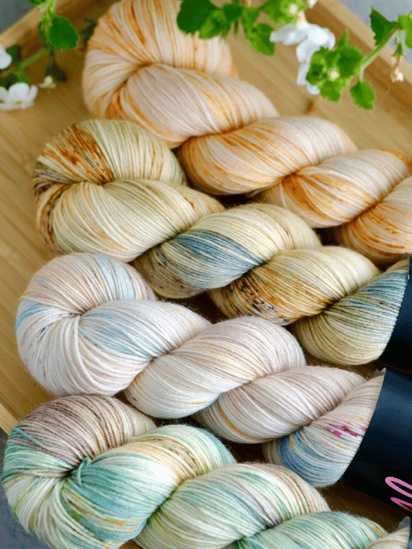 Four skeins of WoolWomen merino sock - Summer Collection yarn in soft cream, gold, blue, and green are arranged on a wooden tray with white flowers in the background.