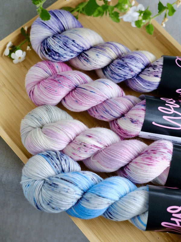 Three skeins of WoolWomen merino sock - Summer Collection yarn in pastel blue, purple, pink, and white are neatly arranged on a wooden tray decorated with small white flowers.