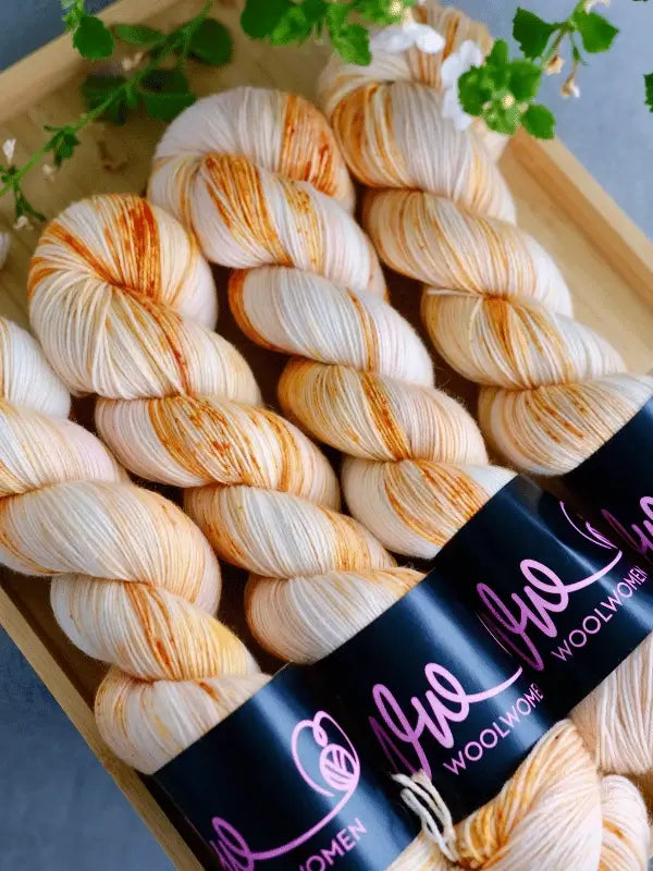 Four skeins of WoolWomen merino sock - Summer Collection yarn, cream-colored with orange speckles and black labels, are neatly displayed in a wooden tray. Green leaves and white flowers decorate the top of the image.
