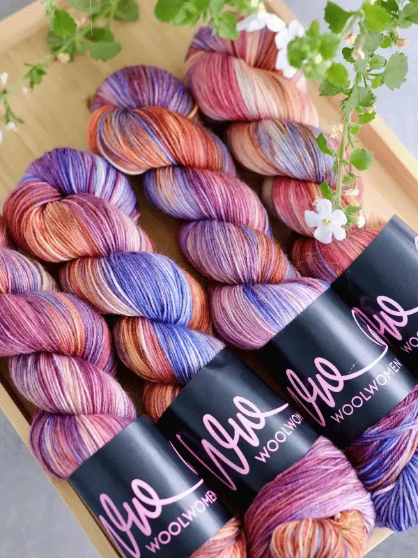 Three skeins of WoolWomen merino sock – Summer Collection yarn in purple, pink, orange, and blue are neatly arranged on a wooden tray with green leaves and white flowers nearby.
