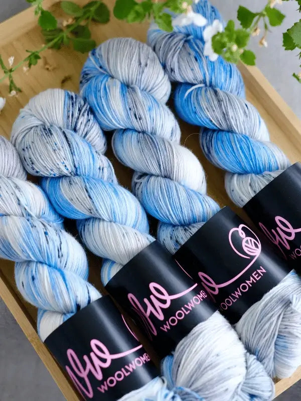 Four skeins of WoolWomen merino sock - Summer Collection yarn, in blue, white, and grey variegated shades with WoolWomen labels, are arranged neatly on a wooden tray with green leaves at the top of the image.