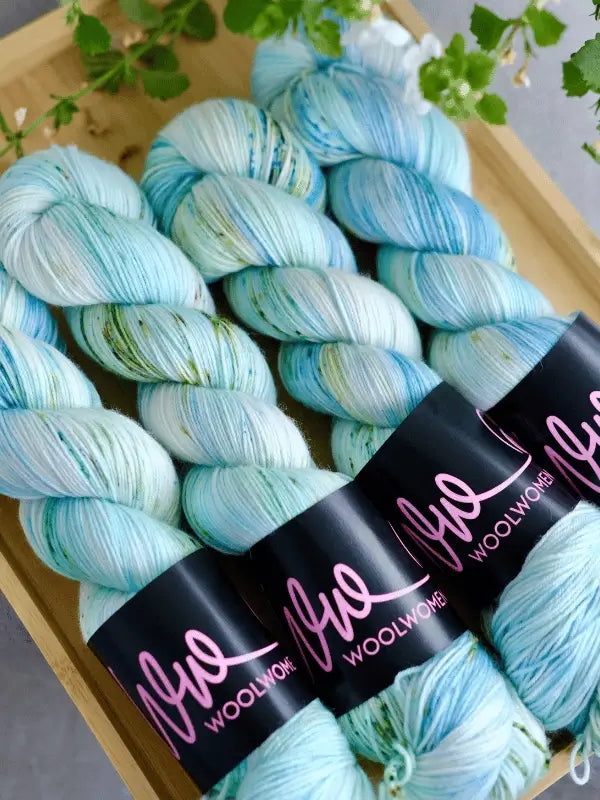Four skeins of blue, white, and mint green speckled WoolWomen merino sock yarn from the Summer Collection are neatly arranged in a wooden tray with green leafy sprigs placed around them.