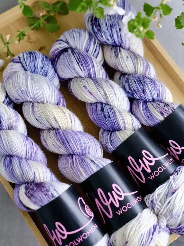 Four skeins of WoolWomen merino sock - Summer Collection yarn in purple and white are neatly arranged on a wooden tray, with green leafy plants nearby.