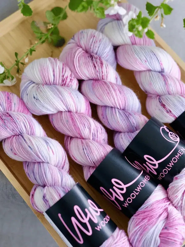 Four skeins of WoolWomen merino sock - Summer Collection yarn in pastel pink, purple, and white sit neatly on a wooden tray with greenery. Each skein features a black “WoolWomen” label.