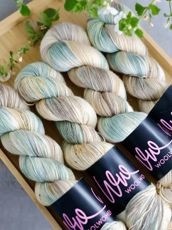 Three skeins of WoolWomen merino sock - Summer Collection yarn in cream, blue, and beige are arranged in a wooden tray with green foliage. Each skein features a black WoolWob label.