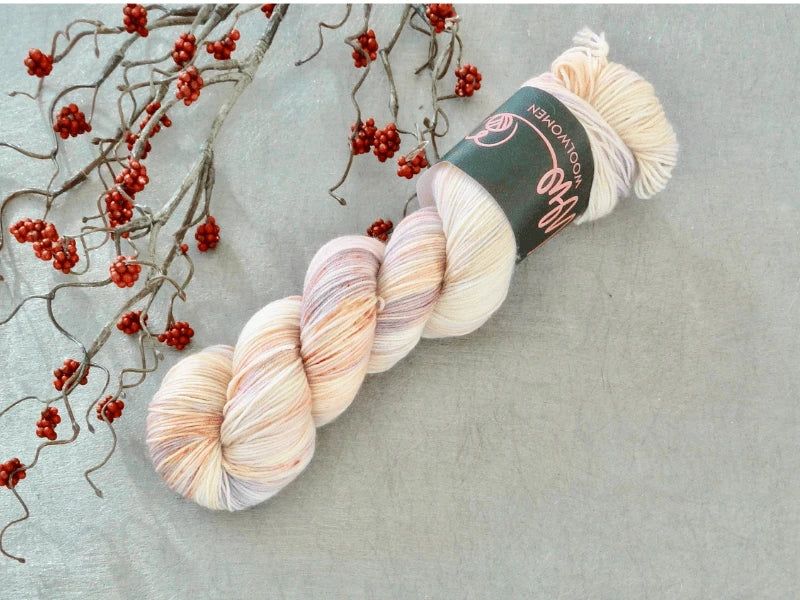 A skein of WoolWomen Merino Sock – Yhden Padan Ihme, a pale multicolored käsinvärjätty lanka, rests on a light grey surface beside a decorative branch with small red berries.