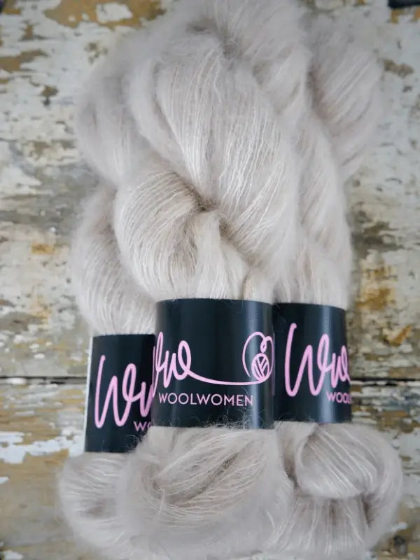 Two skeins of soft, pale grey WoolWomen Silk Mohair yarn with pink labeling rest on a rustic, weathered wooden surface. This käsinvärjätty lanka is a luxurious blend of kid mohair and silk, making it perfect for your next elegant project.