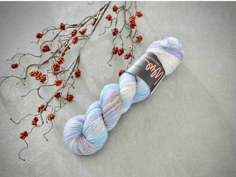 A skein of WoolWomen Merino Sock – Yhden Padan Ihme pastel multicolored yarn rests on a light gray surface beside a branch with small red berries. The label indicates it's mulesing-vapaa merino wool.