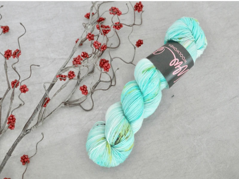 A skein of WoolWomen Merino Sock – Yhden Padan Ihme yarn in turquoise, white, and green rests on a light gray surface beside a dry branch with small red berries.