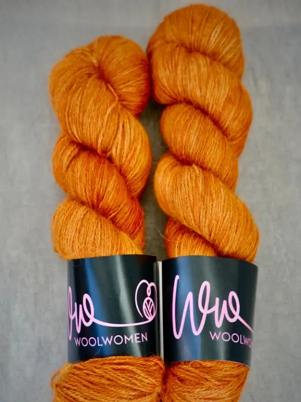 Two skeins of orange WoolWomen AlpaLux baby alpakka yarn are placed side by side on a gray surface. Each skein features a black label with pink writing that reads “Woowoolwomen” alongside a small sheep design, accentuating the luxurious feel and quality of this ylellinen lanka alpakkalanka.