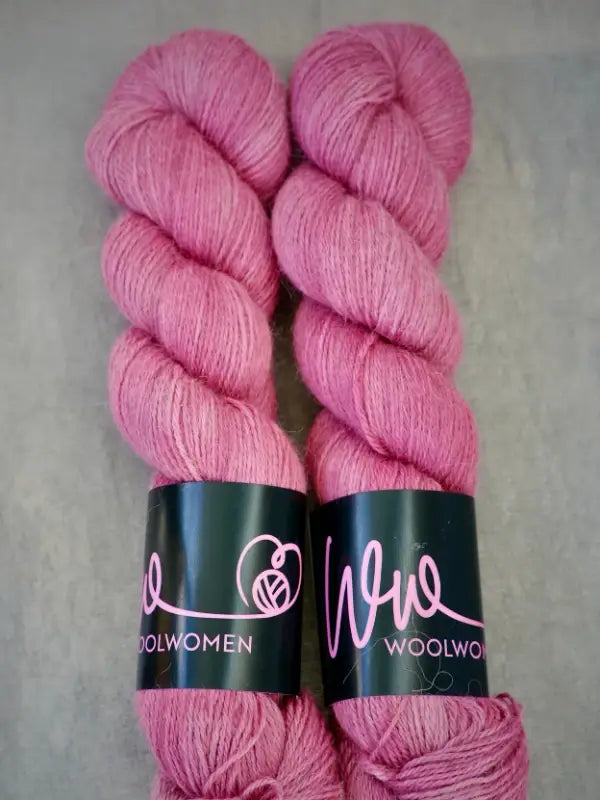 Two skeins of pink WoolWomen AlpaLux baby alpaca yarn are placed side by side on a grey surface, each wrapped with a black label featuring the WoolWomen name and a yarn ball logo.
