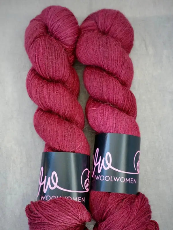 Two skeins of deep red WoolWomen AlpaLux yarn with black labels reading “WoolWomen” and a pink logo, placed side by side on a gray surface.