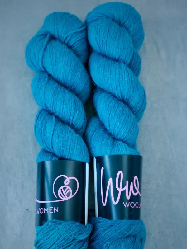 Two skeins of bright teal blue WoolWomen AlpaLux yarn with black labels featuring pink writing and a yarn ball logo, placed side by side on a light gray background.