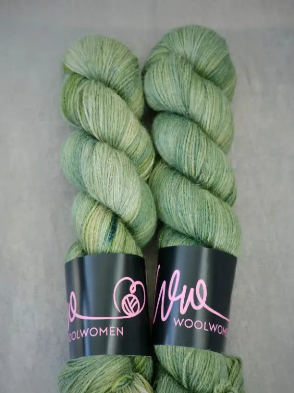 Two skeins of light green WoolWomen AlpaLux yarn, crafted from soft baby alpaca fibers and featuring WoolWomen labels, placed side by side on a gray surface.