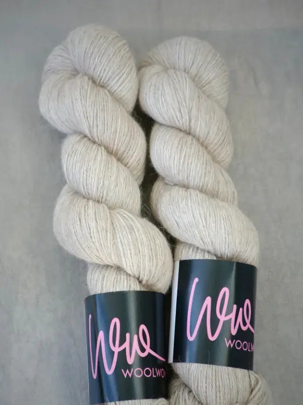 Two skeins of off-white WoolWomen AlpaLux yarn, each with a black label featuring “wool” in pink cursive lettering. The luxuriously soft baby alpakka fibers are neatly twisted and placed side by side on a neutral background.