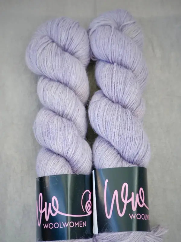Two skeins of pale lavender WoolWomen AlpaLux baby alpaca yarn are placed side by side on a light gray background. Each skein features a black label with pink text that reads WoolWomen, emphasizing this ylellinen lanka’s soft and luxurious feel.