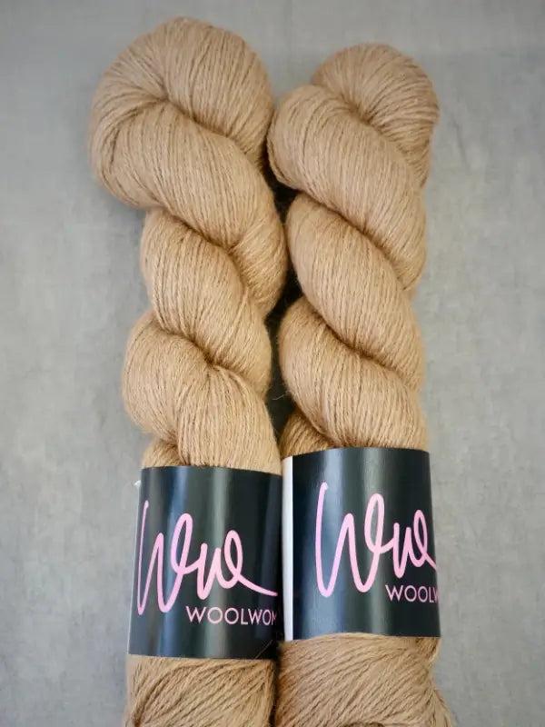 Two skeins of light brown WoolWomen AlpaLux yarn are placed side by side on a gray surface. Each skein has a black label with pink writing that says WoolWomen, emphasizing the ylellinen lanka and baby alpakka softness of this luxurious yarn.