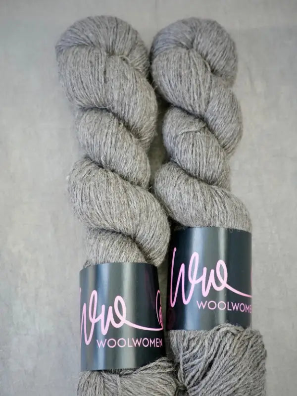 Two skeins of light gray WoolWomen AlpaLux alpakkalanka, each with a black label featuring “Wool Women” in pink script, are placed side by side on a light gray surface. This baby alpakka yarn is perfect for creating soft, ylellinen lanka projects.