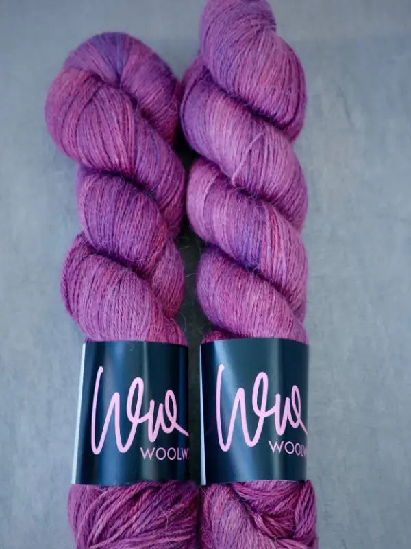 Two skeins of WoolWomen AlpaLux yarn, featuring a luxurious blend of baby alpakka and soft kashmir fibers in purple, lie side by side on a gray background.