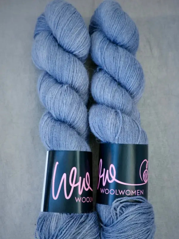 Two skeins of light blue WoolWomen AlpaLux alpakka-lanka yarn with black labels that read “WoolWomen” in pink cursive letters, placed side by side on a gray surface.