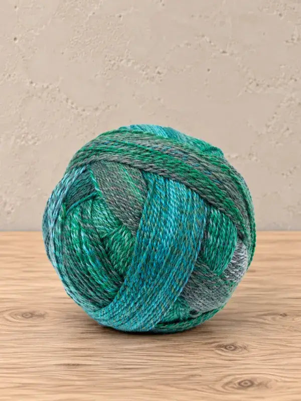 A round ball of Schoppel wolle Zauberball crazy variegated yarn in shades of teal, green, and gray.