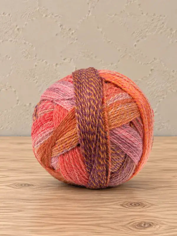 A round ball of Schoppel wolle Zauberball crazy yarn featuring gradients of pink, orange, and purple, with a single darker, marled strand wrapped around the center.