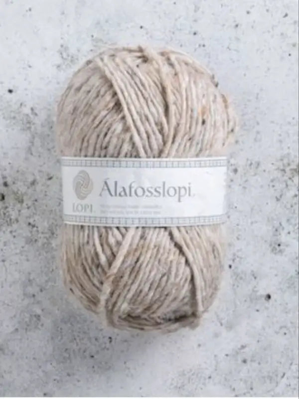A skein of light beige Istex Alafosslopi yarn, renowned for its authentic Icelandic wool, with a white label rests on a light gray, speckled surface.