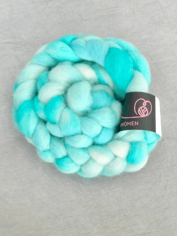 A neatly coiled braid of soft, chunky WoolWomen Suomalainen Kehruuvilla käsinvärjätty in turquoise and white, wrapped with a black label featuring a pink logo and text. The braid rests on a plain light grey background.