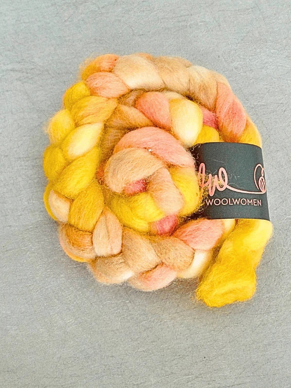 A coiled braid of WoolWomen Suomalainen Kehruuvilla käsinvärjätty in yellow, orange, pink, and beige tones with a black WOOLWOMEN label rests on a light gray surface.
