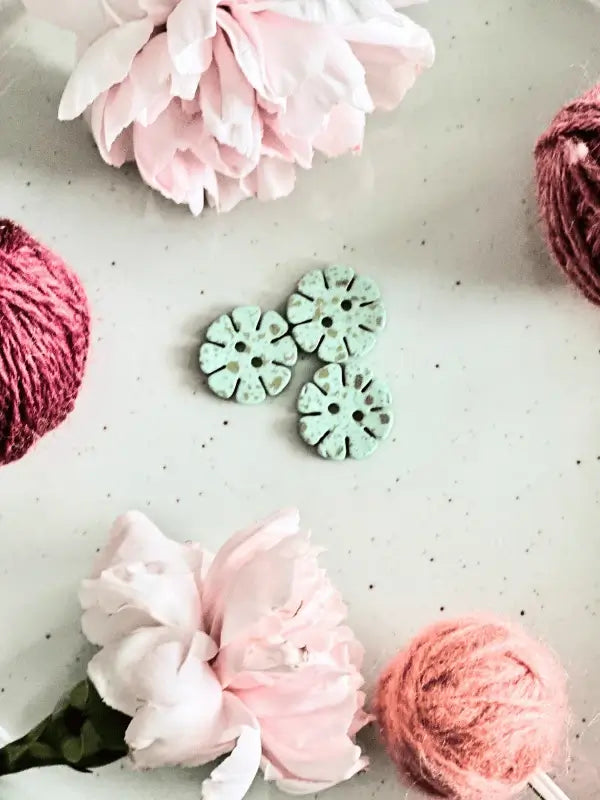 Three Värikkäät kukkanapit 20mm (17700) are centered on a white surface, surrounded by pink peonies and balls of pink and burgundy yarn, creating a simple, charming scene with a natural touch.