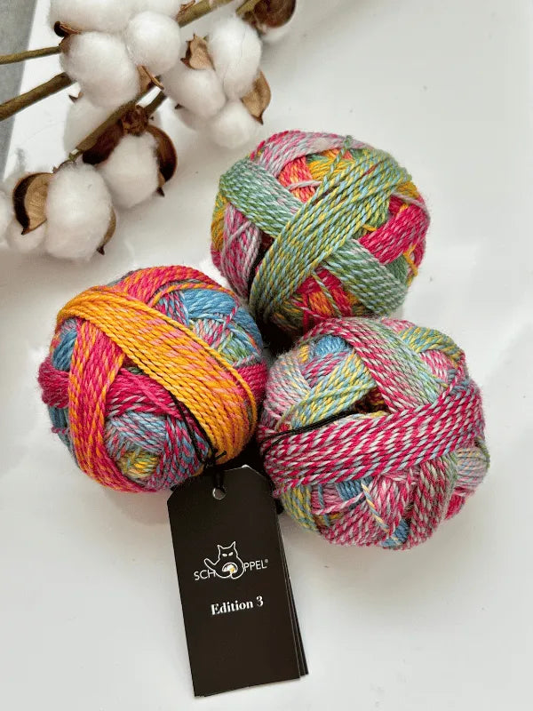 Three vibrant balls of Schoppel-Wolle Edition 3 yarn, featuring blue, pink, yellow, and green hues, are displayed on a white surface beside cotton stems. Each ball bears a black Schoppel Edition 3 tag.