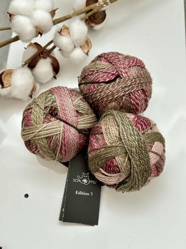 Three balls of Schoppel-Wolle Edition 3 liukuvärjätty lanka in pink, mauve, and olive green rest on a surface beside a cotton branch. A black Edition 3 label is beneath this superwash-merinovilla yarn.
