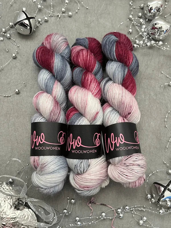 Three skeins of WoolWomen Alpaca Sock - yhden padan ihmeet hand-dyed yarn in gray, pink, and burgundy are arranged on a gray surface with silver holiday decorations. Each soft skein is labeled WoolWomen.