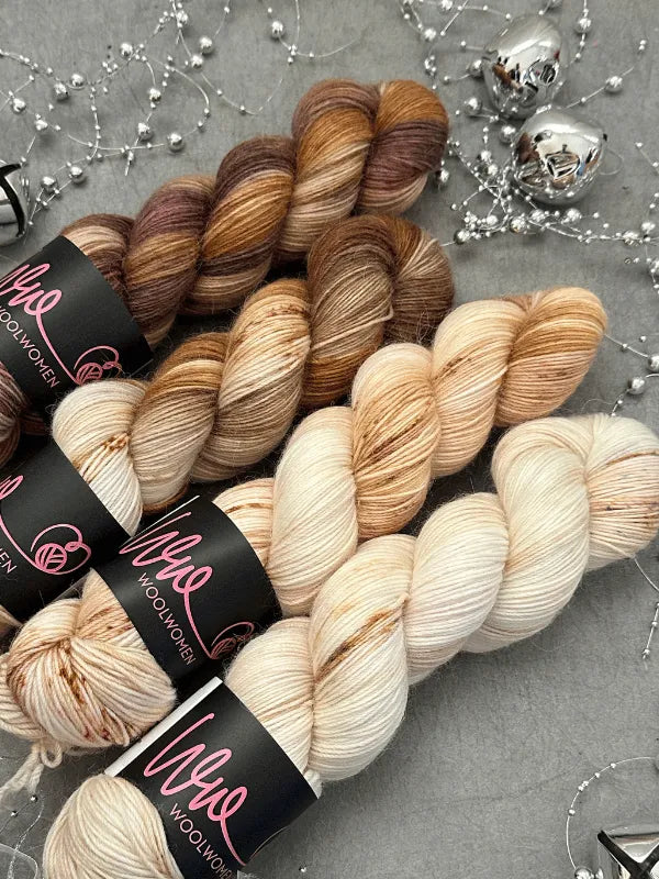 Four skeins of yarn in cream, tan, and brown—including WoolWomen Alpaca Sock - yhden padan ihmeet—are arranged on a gray surface with silver beads and two metallic ornaments nearby.