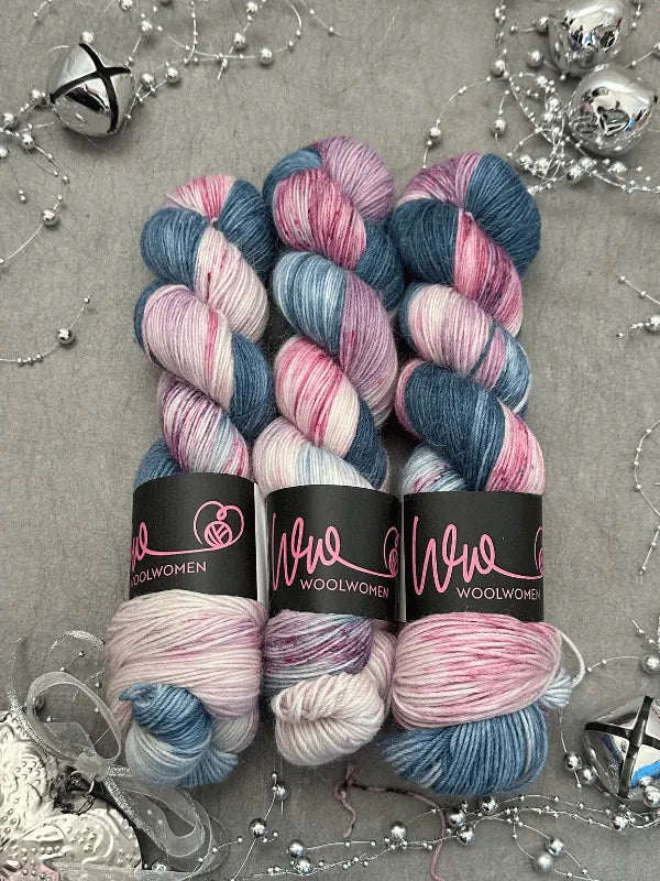 Three skeins of WoolWomen Alpaca Sock - yhden padan ihmeet yarn in white, blue, and pink are arranged on a gray surface with silver holiday decorations, bells, and beads scattered around.