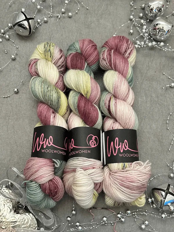 Three skeins of WoolWomen Alpaca Sock - yhden padan ihmeet yarn in soft pink, green, yellow, and cream are arranged on a gray surface with silver beads, ribbon, and decorative bells. Each skein is labeled Wool Women.