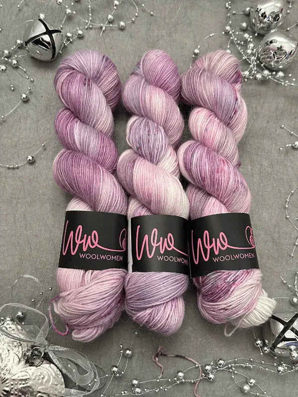 Three skeins of WoolWomen Alpaca Sock -yhden padan ihmeet in pink and lavender variegated tones are displayed on a gray surface, accented with silver beads, jingle bells, and festive decorations—perfect for your next cozy project.