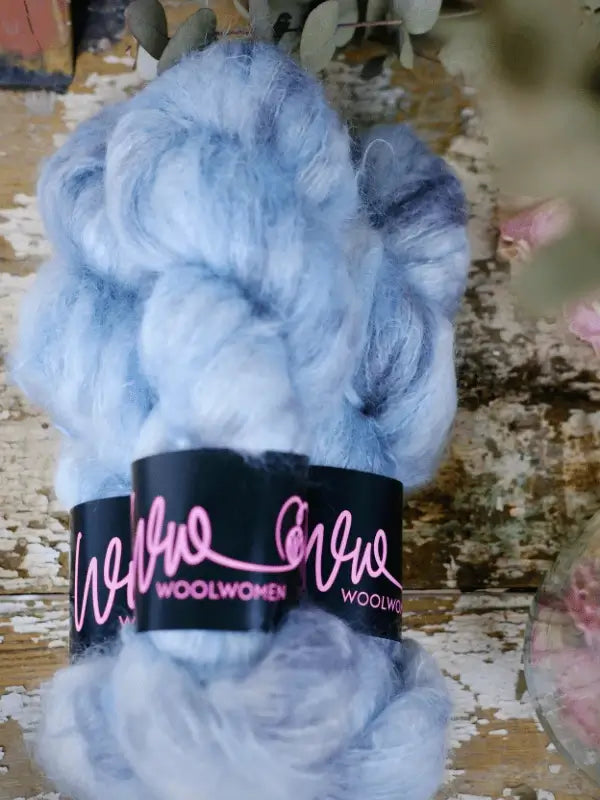 Two skeins of WoolWomen Fluff Mohair yarn in soft, light blue with lavender hints, each wrapped in a black label with pink script, sit on a rustic wooden surface.