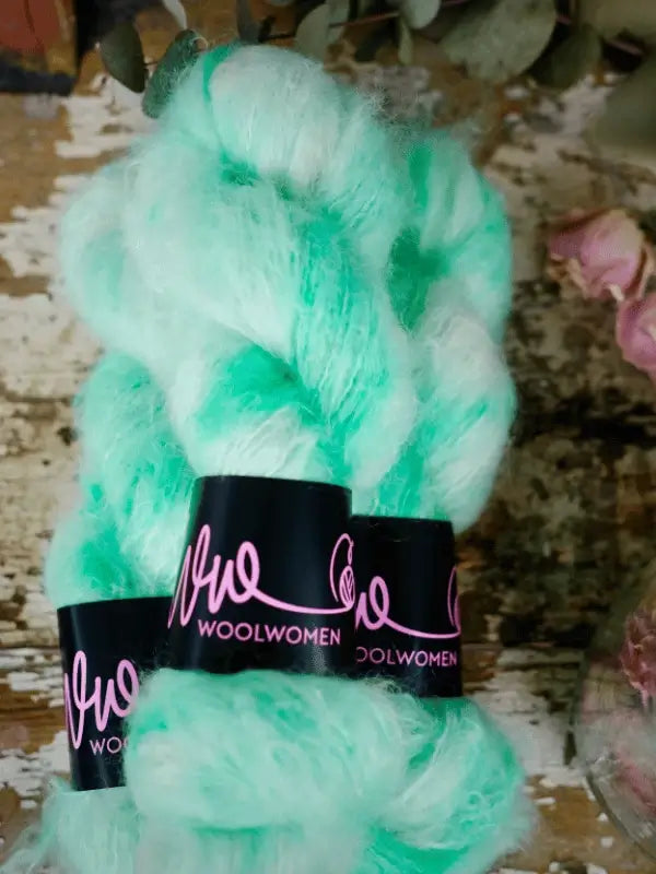 A skein of WoolWomen Fluff Mohair in mint green, featuring a black band with pink lettering, sits on a rustic wooden surface beside dried flowers.
