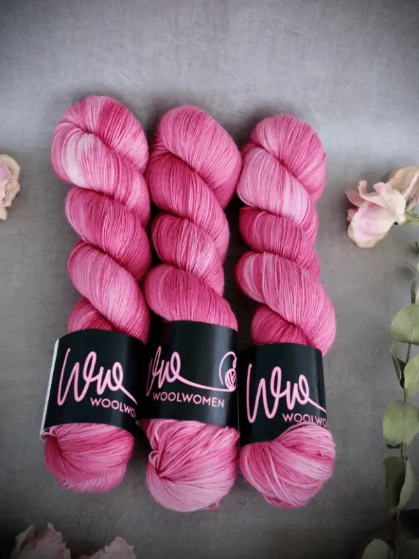 Three skeins of vibrant pink WoolWomen Merino Sock yarn are lined up side by side on a gray surface, surrounded by light pink flowers.