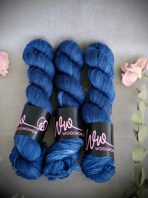 Three skeins of deep blue WoolWomen Merino Sock yarn are arranged side by side on a gray surface, surrounded by pink dried flowers and green leaves.