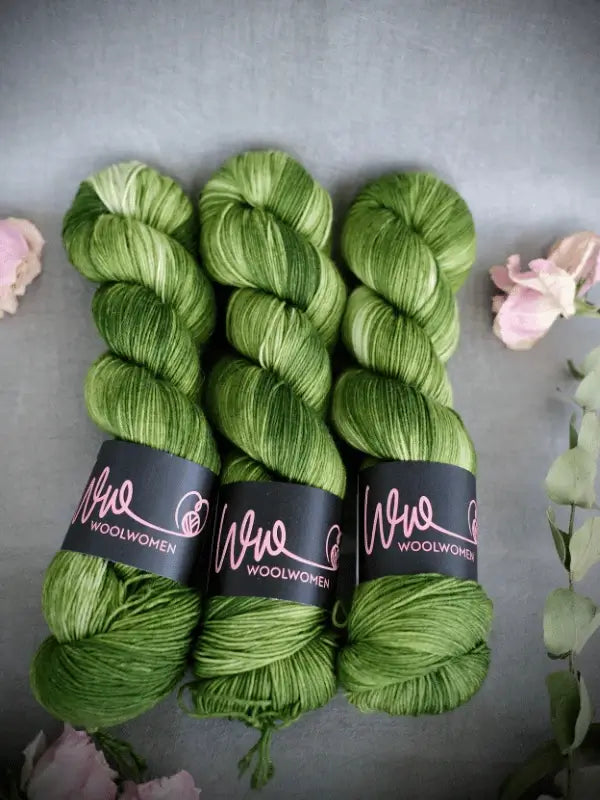 Three skeins of WoolWomen Merino Sock in green tones, made from superwash merino wool, are arranged on a gray surface with black WoolWomen labels and soft pink flowers around them.