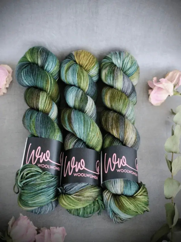 Three skeins of WoolWomen Merino Sock in variegated green, blue, and brown are arranged side by side on a gray surface, surrounded by pink flowers and green leaves.