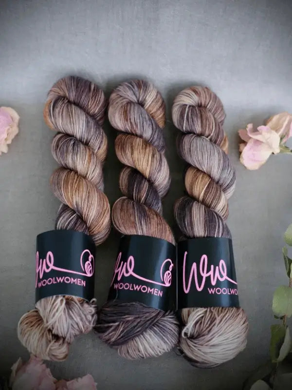Three skeins of brown, tan, and gray variegated WoolWomen Merino Sock yarn are neatly arranged side by side on a gray surface, surrounded by pale pink and white flowers.
