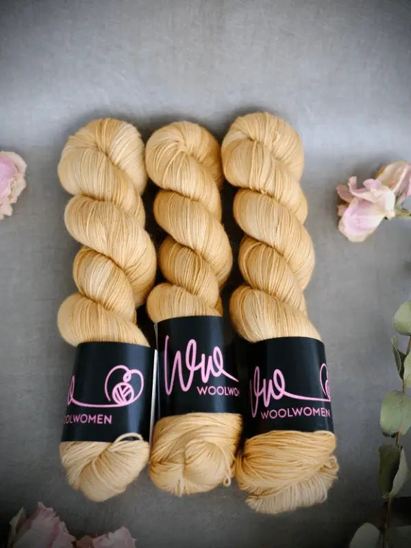 Three skeins of light yellow WoolWomen Merino Sock yarn with black Woolwomen labels are side by side on a gray surface, accented by light pink flowers and green leaves.