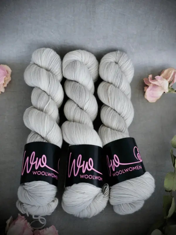 Three skeins of WoolWomen Merino Sock light grey superwash merinovilla yarn with black labels and pink text are arranged side by side on a grey surface, with pink flowers in the corners of the image.