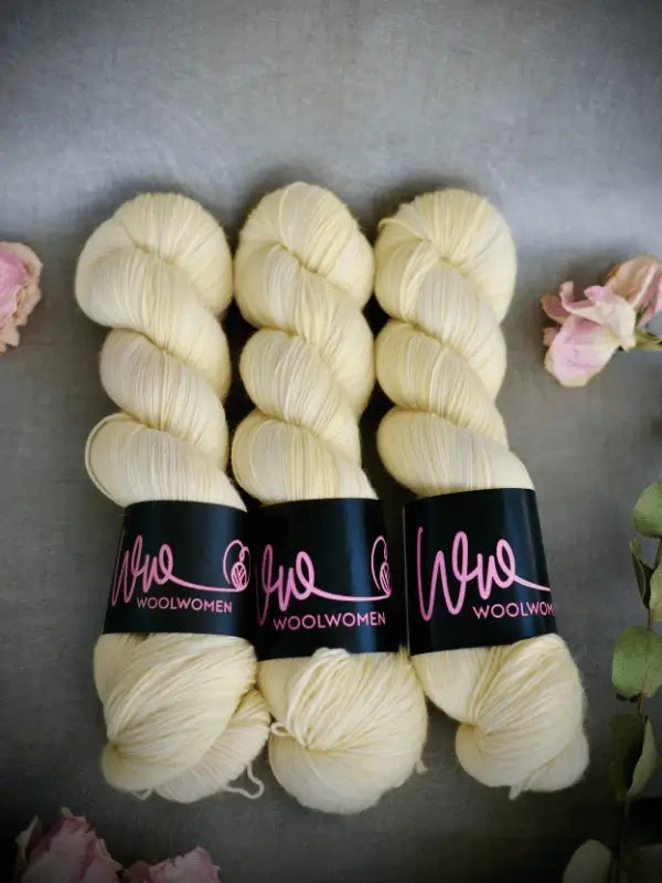 Three skeins of pale yellow WoolWomen Merino Sock yarn with black and pink labels are arranged side by side on a gray surface, surrounded by dried pink flowers and green leaves.