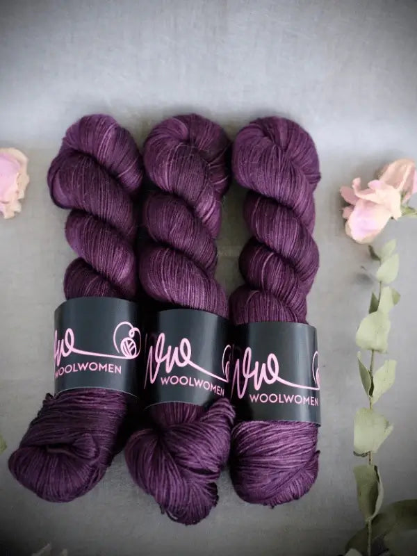 Three skeins of deep purple WoolWomen Merino Sock yarn are arranged side by side on a gray surface, surrounded by light pink flowers and eucalyptus leaves.