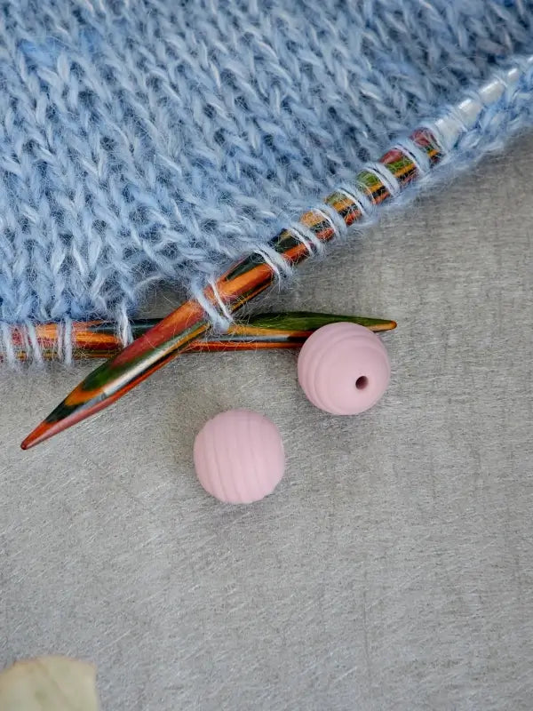 Woolwomen Puikkostopparit knitting needle stoppers in ribbed pink rest alongside colorful knitting needles on light blue knitted fabric, all arranged on a light gray surface.